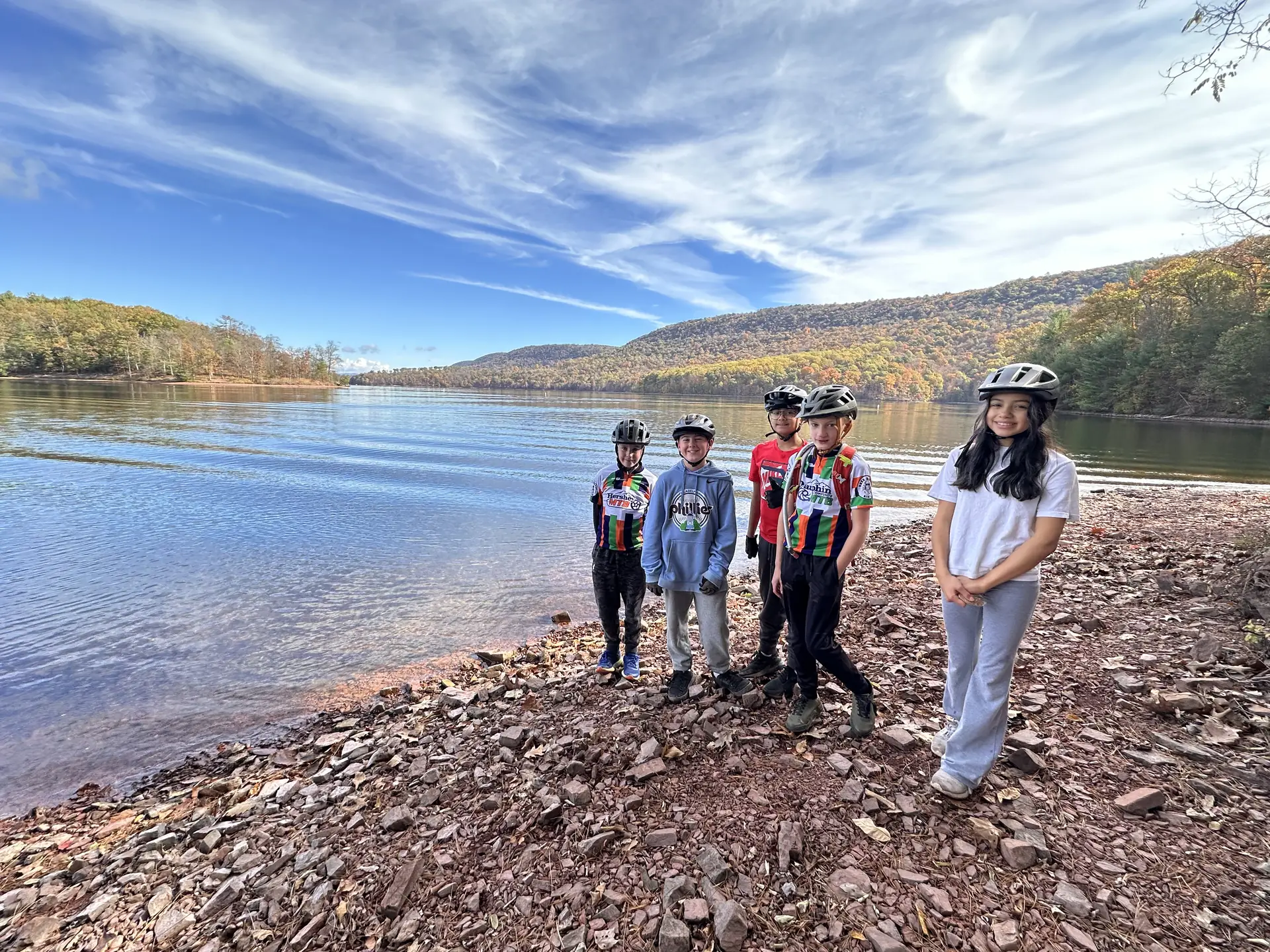 Five helmeted teammates smile beside Raystown Lake with autumn hills reflected in the water.